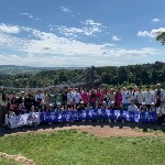 a group of students posing for a photo on a sunny day with a bridge behind them